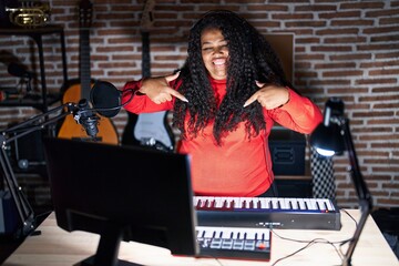 Plus size hispanic woman playing piano at music studio looking confident with smile on face, pointing oneself with fingers proud and happy.