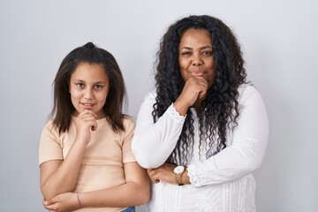 Mother and young daughter standing over white background with hand on chin thinking about question, pensive expression. smiling and thoughtful face. doubt concept.