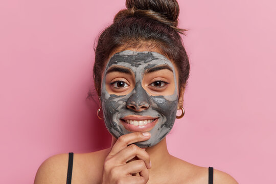 Portrait of lovely Indian woman keeps hand on chin and smiles gently focused at camera has dark eyes applies nourishing clay mask for skin exfoliating and deep cleansing isolated over pink background.