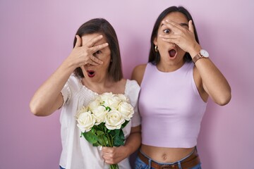 Hispanic mother and daughter holding bouquet of white flowers peeking in shock covering face and eyes with hand, looking through fingers with embarrassed expression.