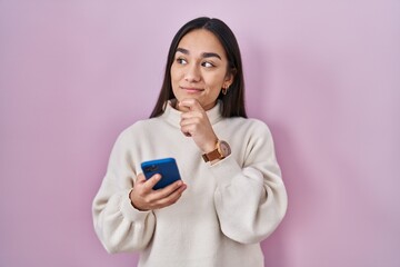 Young south asian woman using smartphone serious face thinking about question with hand on chin, thoughtful about confusing idea - obrazy, fototapety, plakaty