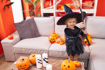 Adorable blonde girl wearing witch costume holding broom at home