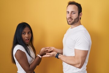 Interracial couple standing over yellow background pointing aside with hands open palms showing copy space, presenting advertisement smiling excited happy