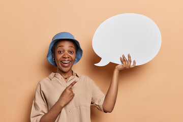 Hey look at this. Glad dark skinned girl points index finger at white speech bubble with empty space for your advertising content smiles pleasantly wears blue panama shirt stands against brown wall