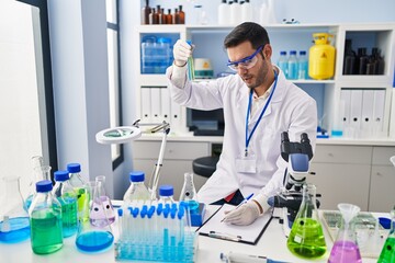 Fototapeta premium Young hispanic man scientist holding test tubes writing report at laboratory