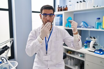 Young hispanic man with beard working at scientist laboratory holding blue ribbon covering mouth with hand, shocked and afraid for mistake. surprised expression