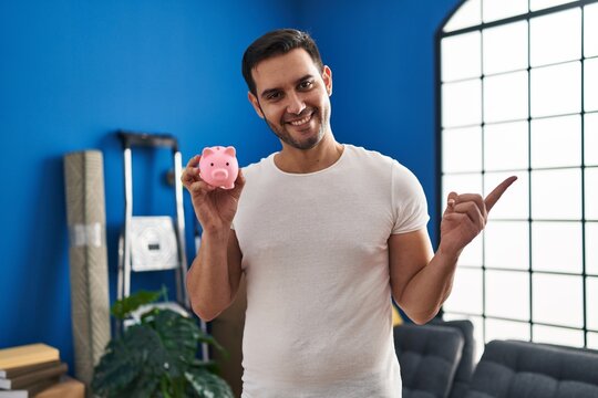 Young hispanic man with beard holding piggy bank at new home smiling happy pointing with hand and finger to the side