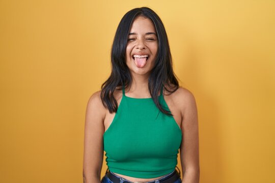 Brunette woman standing over yellow background sticking tongue out happy with funny expression. emotion concept.
