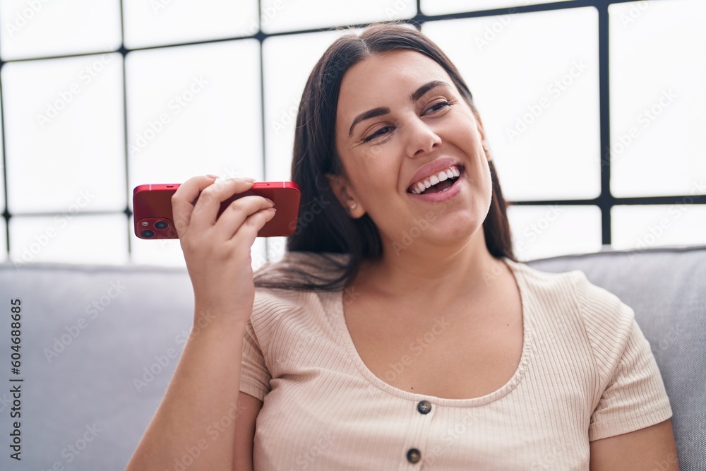 Canvas Prints Young beautiful hispanic woman listening voice message by smartphone sitting on sofa at home