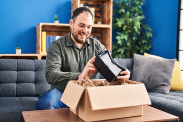 Young hispanic man unpacking virtual reality glasses sitting on sofa at home