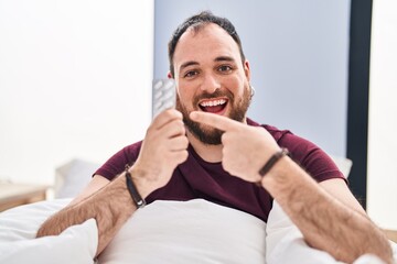 Plus size hispanic man with beard in the bed holding pills smiling happy pointing with hand and finger