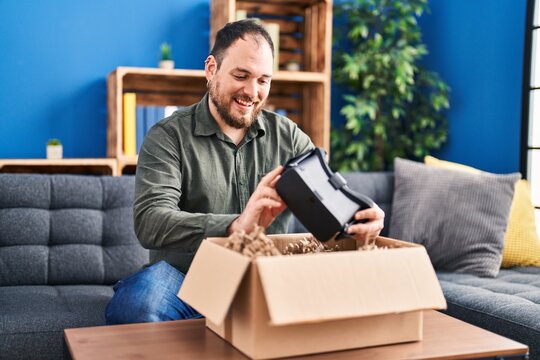 Young hispanic man unpacking virtual reality glasses sitting on sofa at home
