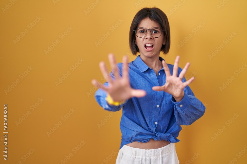 Wall mural Young girl standing over yellow background afraid and terrified with fear expression stop gesture with hands, shouting in shock. panic concept.