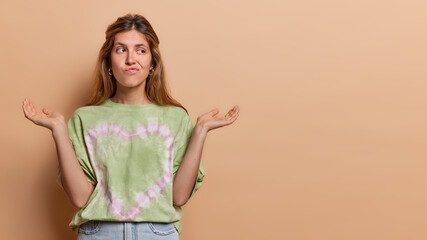 Obraz premium Studio shot of confused woman purses lips and spreads palms feels questioned cannot finds right decision dressed in casual clothes isolated over brown background copy space for your advertisement