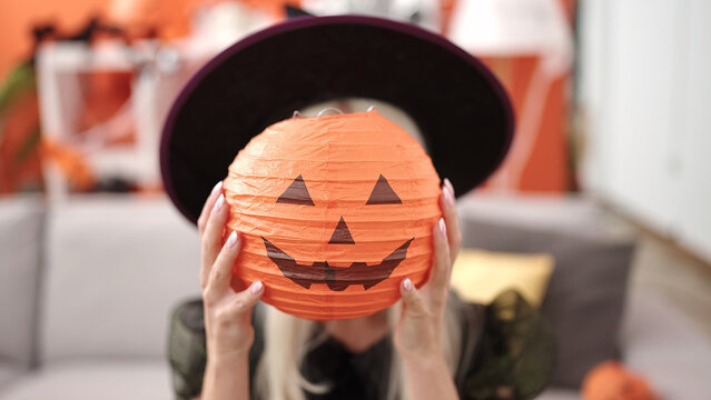 Young blonde woman wearing witch costume holding halloween pumpkin basket over face at home