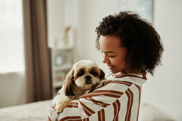 Minimal side view portrait of black young woman holding cute pet dog at home and smiling