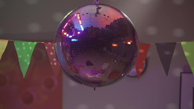 A silvery mirrored disco ball rotates and throws multicolored highlights on the walls close up. Party with disco ball and paper flags. Party, birthday, celebration, disco.HDR BT2020 HLG Material.
