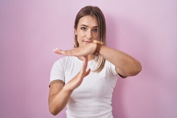 Blonde caucasian woman standing over pink background doing time out gesture with hands, frustrated and serious face