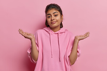 Clueless indecisive Indian woman spreads palm shrugs shoulders looks clueless at camera dressed in casual sweatshirt cannot decide or answer your question isolated over pink background faces dilemma - obrazy, fototapety, plakaty