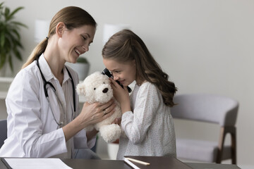 Cute little girl, clinic patient having fun, holds otoscope tool, play otolaryngologist, check ear of fluffy toy bear, during visit in hospital with friendly medical worker. Childcare, otolaryngology - obrazy, fototapety, plakaty