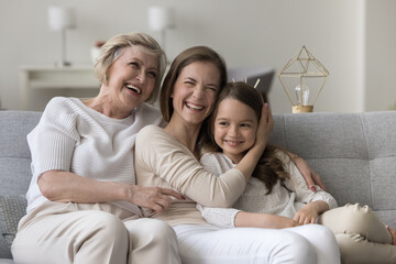 Happy elderly 60s woman, her adult daughter and cute little grandchild laughing, joking seated on...