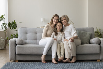 Three generations of women sit on sofa smile look at camera. Elderly 60s grandma her cute little grandchild and grownup daughter shooting for picture in living room. Multigenerational family portrait