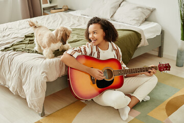 Candid portrait of black young girl playing acoustic guitar and relaxing at home with little pet dog, copy space