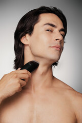 Confident young shirtless man using electric razor while shaving against grey background