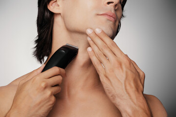 Close-up of unrecognizable shirtless man using electric razor while shaving against grey background