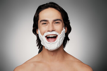 Handsome young shirtless man with shaving cream on face smiling against grey background