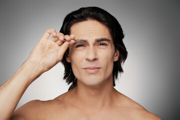Fototapeta premium Handsome young shirtless man plucking his eyebrows against grey background