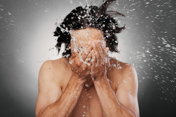 Unrecognizable shirtless man washing face with water splashing around against grey background