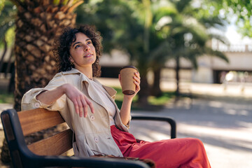 Pretty serene young woman with cup of coffee to go sitting on park bench in city downtown, looking...