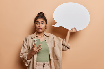 Studio shot of impressed Indian woman holds smartphone in one hand and blank speech bubble in other reacts to something surprising suggests to place your promotional text wears shirt and trousers