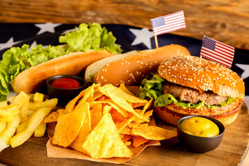 Wooden board with tasty hot dogs, burger, nachos and french fries, closeup. Memorial Day celebration