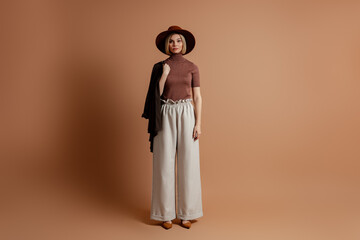 Full length of fashionable young woman in elegant hat standing against brown background