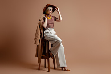 Fashionable young woman in elegant hat sitting on a chair against brown background