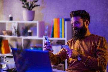 A happy smart casual bearded hipster is sitting at a neon blue lighted home office and using a cellphone and credit card for online purchases.