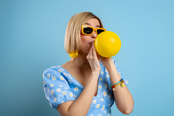 Playful young woman blowing up a balloon while standing against blue background