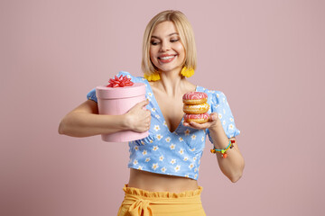 Happy young woman holding gift box and doughnuts against pink background