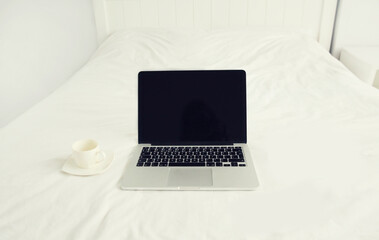 Laptop with blank black screen and cup of coffee on white bed in hotel room