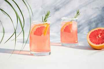 Two glasses with a fresh, cold summer cocktail of grapefruit and ice, garnished with a sprig of rosemary and fresh fruit slices. Red orange drink. Shadows on a sunny day.