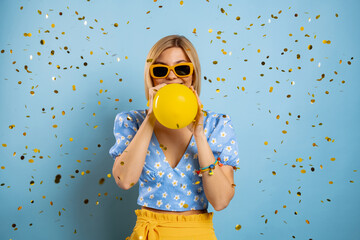 Happy young woman blowing up a balloon while confetti flying around against blue background