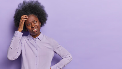 Puzzled dark skinned woman scratches head and looks doubtful aside considers options cannot make decision dressed in purple shirt poses indoor blank copy space for yoour advertising content.