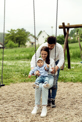 happy mom with dad and small child ride on a swing. young parents on the playground with a little boy. dad rides mom and baby on a swing