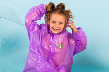 Charming little girl in a purple raincoat puts on a hood. A child dressed for a rainy weather. Isolated on blue background.