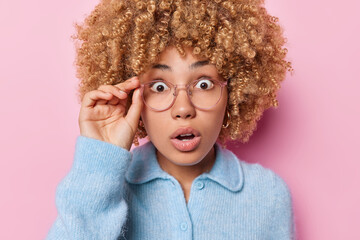 Portrait of shocked emotional curly haired woman stares through spectacles gazes with stupefaction hears surprising news dressed in blue sweater isolated over pink background. Human reactions concept - obrazy, fototapety, plakaty