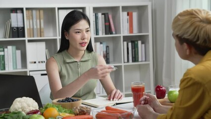 Woman Asian Professional Nutritionist explaining the data on the phone to her client, surrounded by a variety of fruits, nuts, vegetables, and dietary supplements on the table - Powered by Adobe