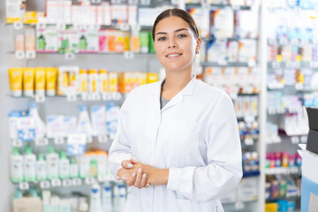 In pharmacy, positive female chemist stands and waits for visitors. Wide range of prescription and over-counter medications, patient care products, baby food