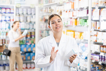 In cosmetic department of pharmacy, female specialist makes consultation about new serum from manufacturer of beauty care products. Client shopping in background